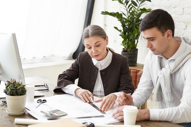 Team of two colleagues interior designers working on architectural project in modern office. Candid shot of young male and mature female architects discussing set of blueprints spread out on desk