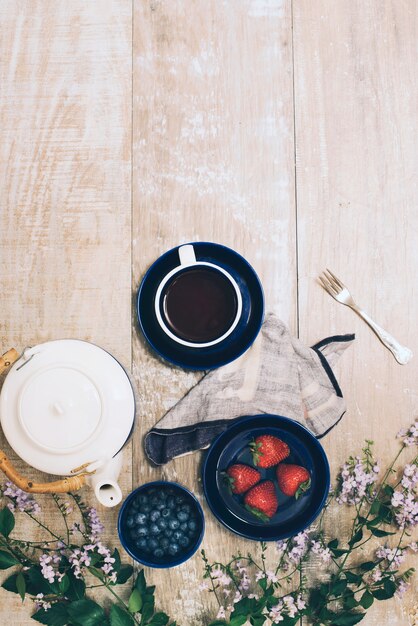 Teapot; coffee cup; berries; fork with napkin and flowers on wooden textured backdrop