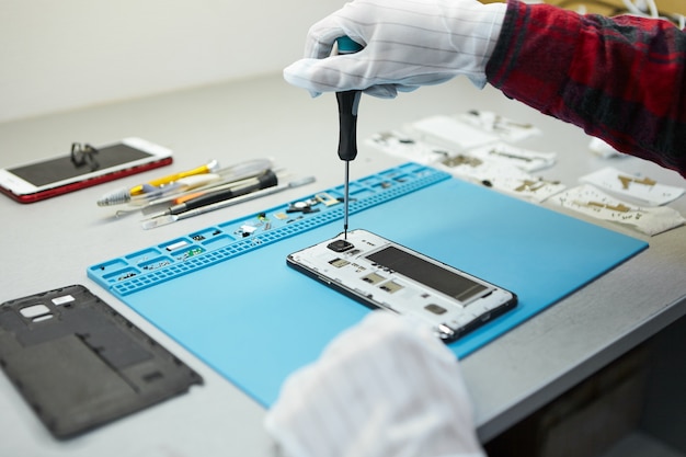 Free photo technician in antistatic gloves using screwdriver to disassemble borken mobile phone, going to repair motherboard, sitting at his workplace in laboratory with needed equipment