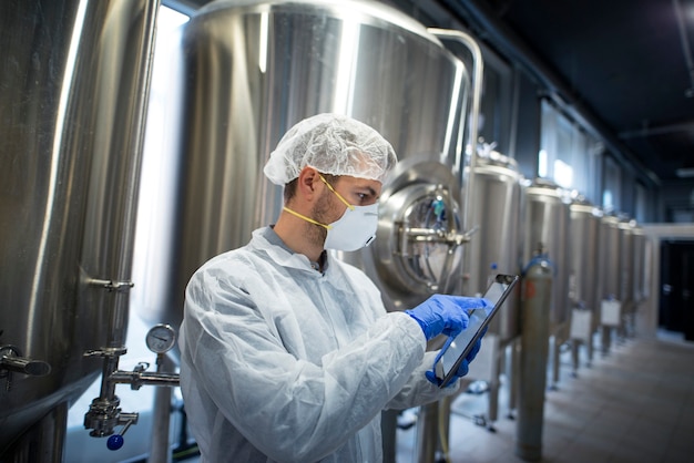 Technologist in protective suit holding tablet and checking production in factory plant