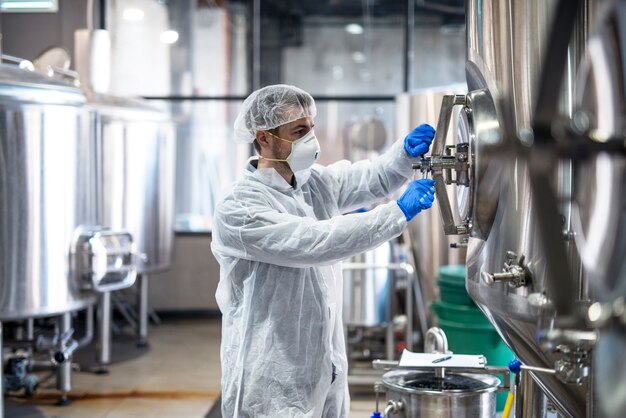 Technologist in protective uniform wearing hairnet mask goggles and gloves closing industrial tank reservoir