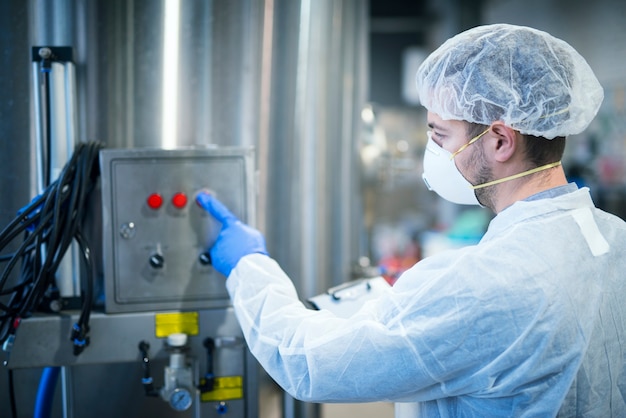 Technologist in white protective uniform with hairnet and mask operating on industrial machine for food processing