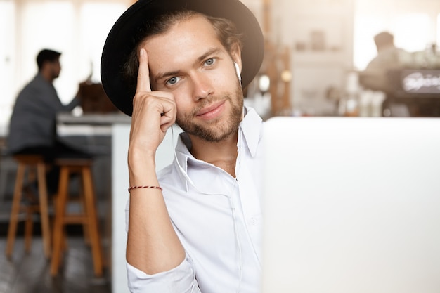 Free photo technology and communication concept. young stylish man with beard wearing black hat listening to music on white earphones sitting in front of generic laptop computer, leaning on his elbow and smiling