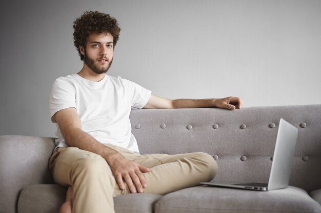 Technology, electronic gadgets and communication concept. Stylish young man wing thick beard and wavy hair sitting on couch in front of open portable computer, using wireless internet connection