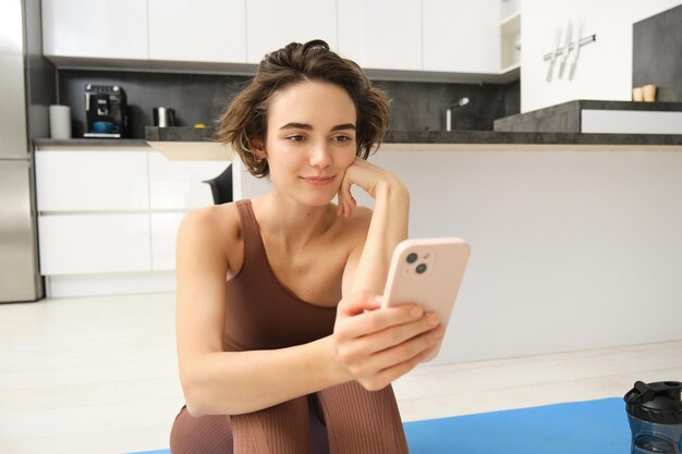 Free photo technology and sport concept fitness girl looking at her smartphone sitting in workout clothes at ho