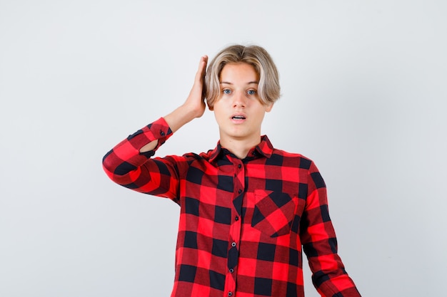 Teen blond male in casual shirt keeping hand near ear and looking pensive , front view.