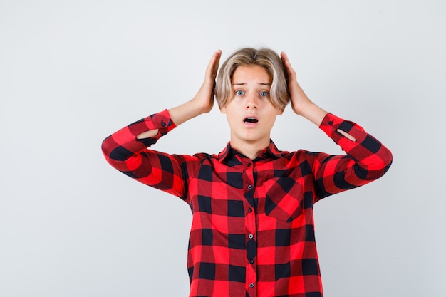 Teen blond male in casual shirt with hands near head and looking scared , front view.