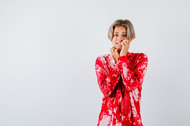 Teen blonde male biting his nails in oversized shirt and looking stressed , front view.