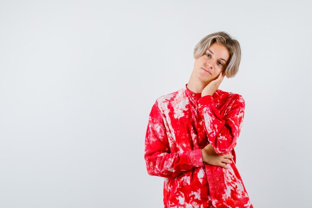 Teen blonde male leaning cheek on palm in oversize shirt and looking pensive. front view.