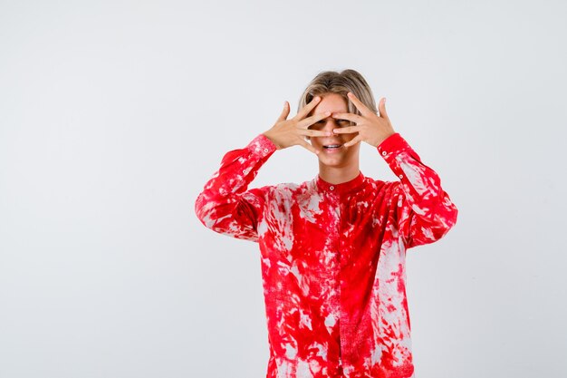 Teen blonde male looking through fingers in oversized shirt and looking curious , front view.