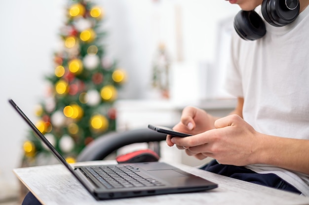 Teen boy is using smartphone with headphones, laptop on the knees at home. Christmas tree on the wall