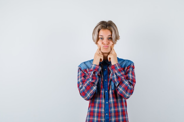 Teen boy pressing fingers on cheeks in checkered shirt and looking disappointed. front view.