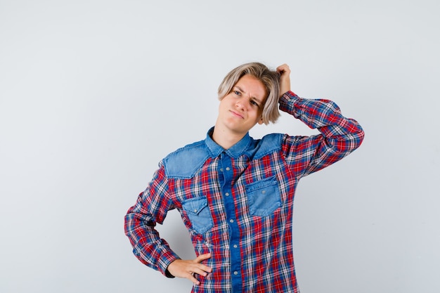 Teen boy scratching head while looking aside in checkered shirt and looking thoughtful. front view.