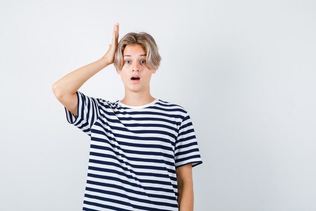 Teen boy in t-shirt keeping hand on head while opening mouth and looking shocked , front view.