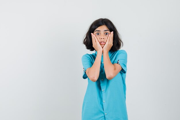 Teen girl in blue shirt holding hands on her cheeks and looking surprised