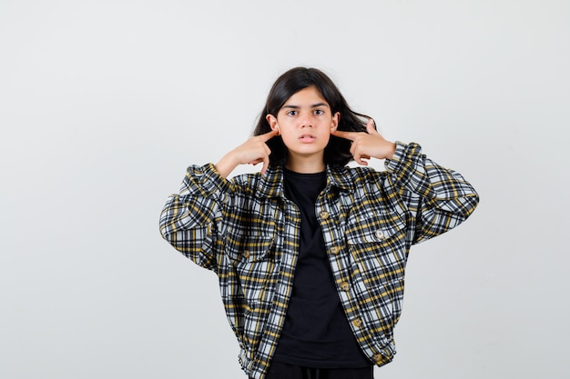 Teen girl in casual shirt touching earlobes with fingers and looking cheerless , front view.