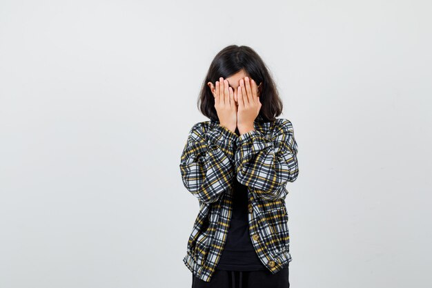 Teen girl covering face with hands in casual shirt and looking annoyed , front view.