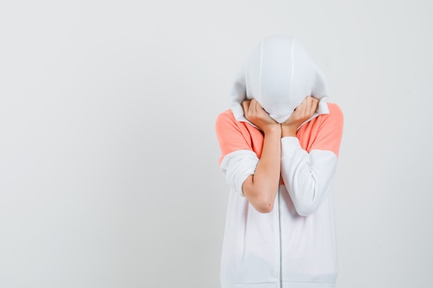 Free photo teen girl covering her head and face with cap in white jacket and looking hidden.