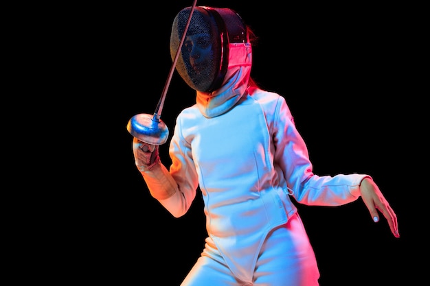 Teen girl in fencing costume with sword in hand isolated on black background, neon light.