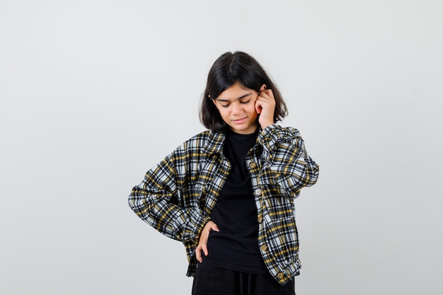 Teen girl holding hand on face, keeping hand on waist in casual shirt and looking cheerful. front view.