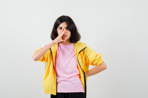 Teen girl holding hand on face, keeping hand on waist in t-shirt, jacket and looking pensive , front view.