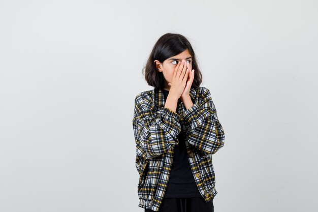 Teen girl holding hands on face, looking aside in casual shirt and looking shocked , front view.