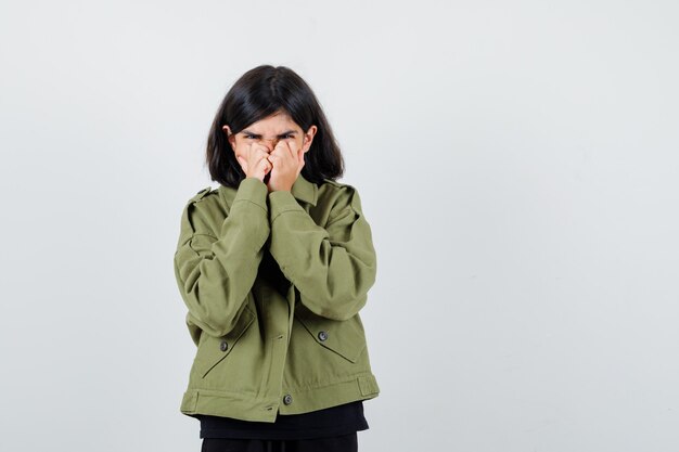 Teen girl holding hands on mouth in t-shirt, green jacket and looking confused. front view.