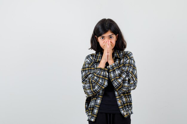 Teen girl holding hands in praying gesture in casual shirt and looking focused , front view.