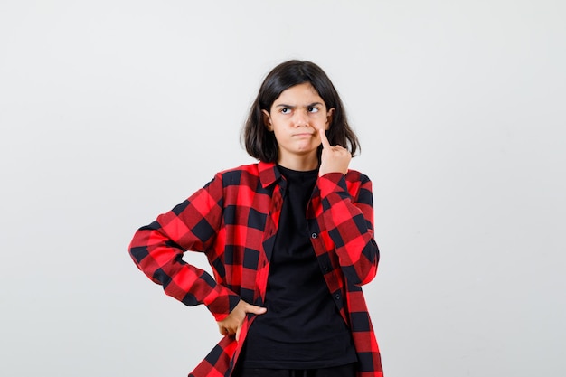 Teen girl holding little finger on cheek in t-shirt, checkered shirt and looking dissatisfied , front view.