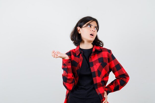 Teen girl posing while looking aside, spreading palm aside in t-shirt, checkered shirt and looking perplexed , front view.