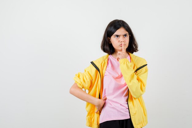 Teen girl in t-shirt, jacket showing silence gesture, looking away and looking focused , front view.