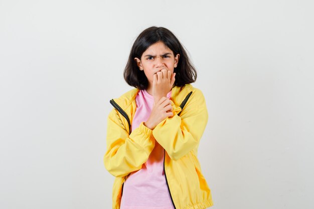 Teen girl in t-shirt, yellow jacket biting nails and looking stressed , front view.