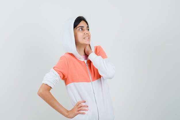 Teen girl in white jacket holding hand on her neck while looking aside and looking nervous.