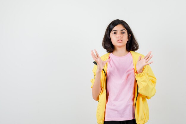 Teen girl in yellow tracksuit, t-shirt spreading palms and looking shocked , front view.