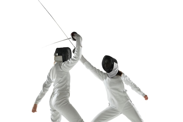 Free photo teen girls in fencing costumes with swords in hands isolated on white studio background. young female models practicing and training in motion, action. copyspace.