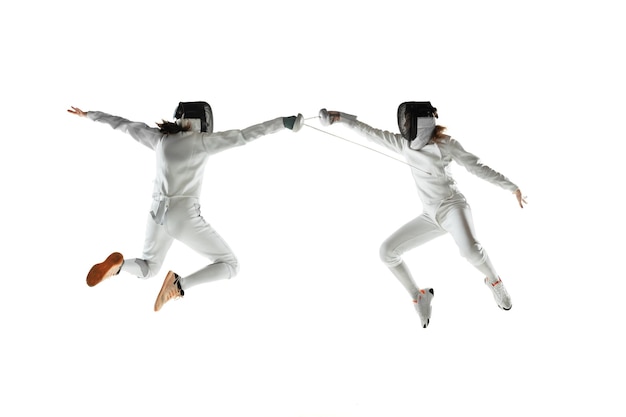 Free photo teen girls in fencing costumes with swords in hands isolated on white studio background. young female models practicing and training in motion, action. copyspace.