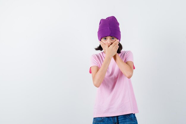 Teen woman holding hands on mouth in t-shirt and beanie looking scared