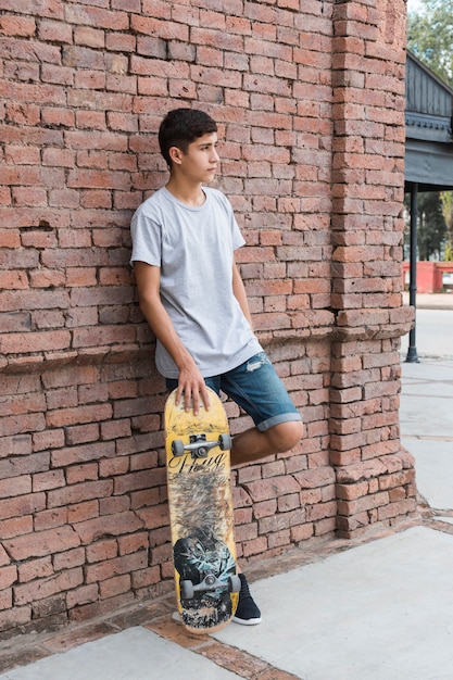 Teenage boy leaning on brick wall holding skateboarding looking away