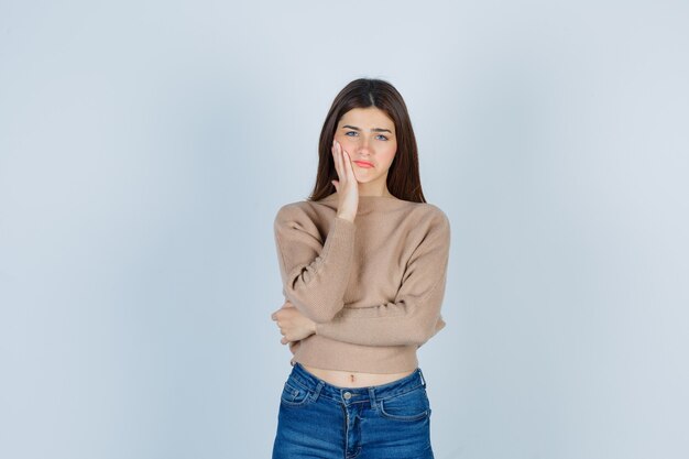 Teenage girl leaning cheek on hand in sweater, jeans and looking upset. front view.