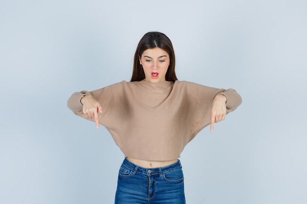 Free photo teenage girl pointing down in sweater, jeans and looking amazed. front view.