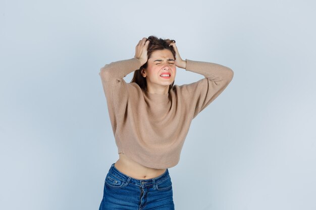 Free photo teenage girl in sweater, jeans clasping head with hands and looking irritated , front view.