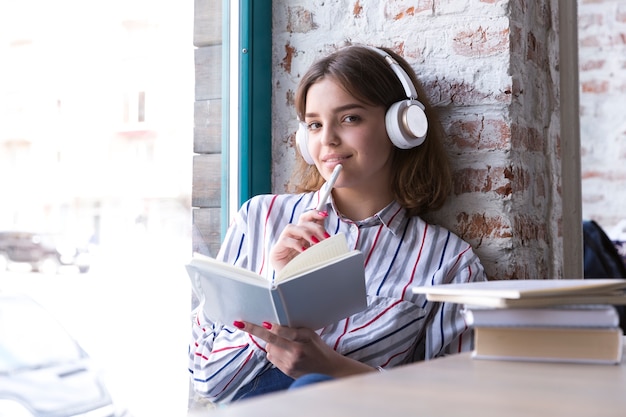 Teenager girl in headphones sitting with open book and looking at camera