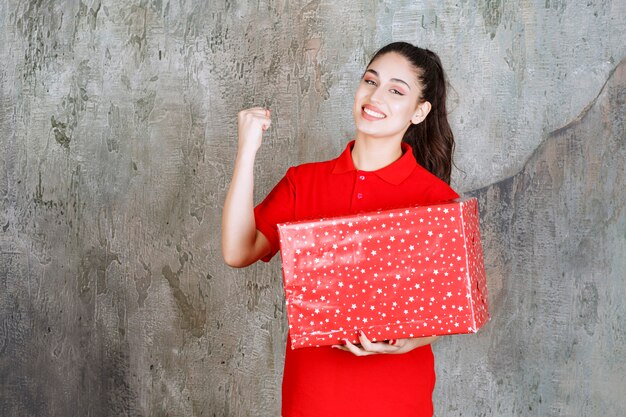 Teenager girl holding a red gift box with white dots on it and showing her fist