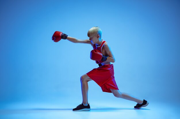 Teenager in sportswear boxing isolated on blue studio background in neon light. Novice male caucasian boxer training hard and working out, kicking. Sport, healthy lifestyle, movement concept.