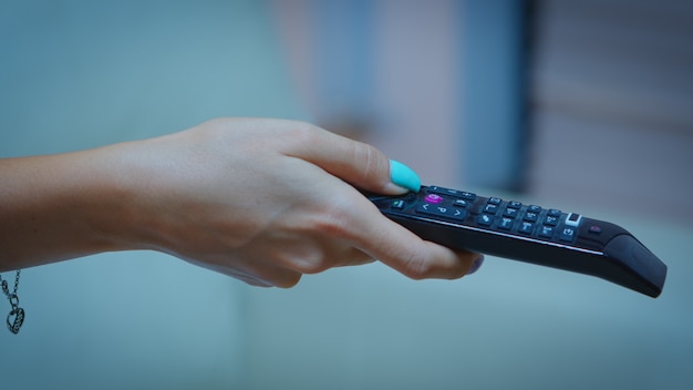 Television remote control in the hands of woman pointing to TV and changing channels. Close up of woman holding controller and pressing the button sitting on couch in front of television.