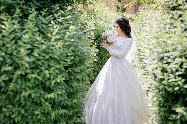 Tender brunette bride is standing in the green garden and sniff wedding bouquet scent