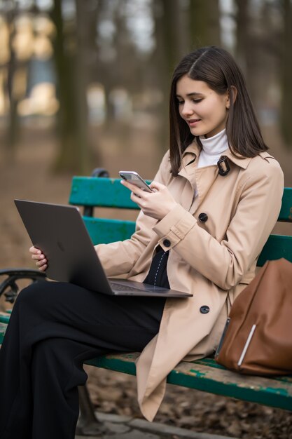 Tender brunette model girl works on laptop and phone outside in the park