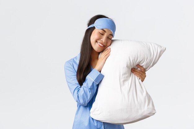 Tender dreamy beautiful asian girl in blue pajamas and sleeping mask lying in bed with closed eyes and hugging pillow, smiling carefree as having good night sleep, standing white background.