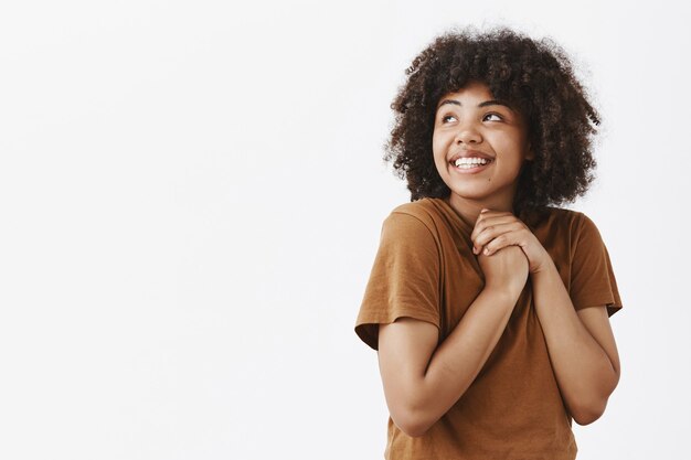 tender and dreamy cute dark-skinned curly-haired woman in brown trendy t-shirt holding hands clenched on chest and gazing at upper left corner with affection