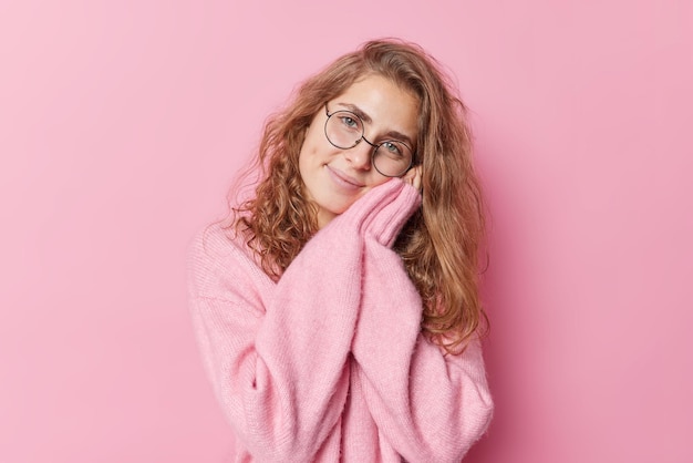 Tender gentle young European woman keeps hands near face smiles happily wears round spectacles and cashmere jumper isolated over pink background. Smiling long haired female model gazes at camera